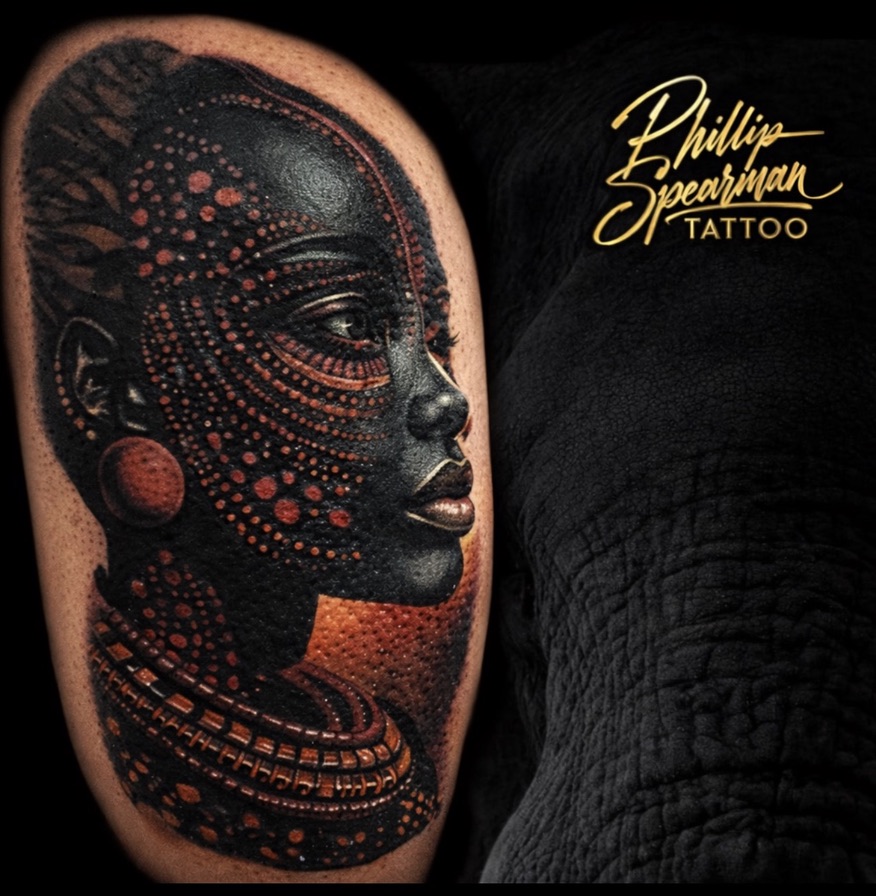 This striking color portrait beautifully captures an African woman's heritage, with deep black ink providing a stunning contrast to the vibrant, tactile reddish-orange patterns. The delicate dots and smooth tonal transitions bring her graceful profile to life, softly illuminated by the interplay of light and shadow. It's a powerful statement of identity, worn with quiet strength on the arm.