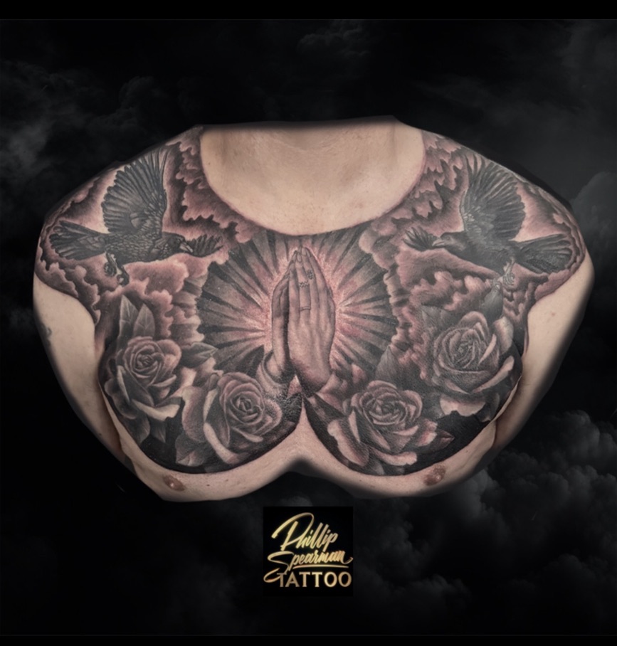 This expansive black and grey chest piece unfolds a deeply personal narrative, with the ethereal glow around the praying hands at its heart, complemented by the delicate texture visible in each rose petal. Above, the dramatic presence of two crows is set against clouds, their smooth tonal transitions creating a seamless and powerful backdrop. This kind of placement for such profound imagery speaks to a permanent dedication of faith or remembrance.
