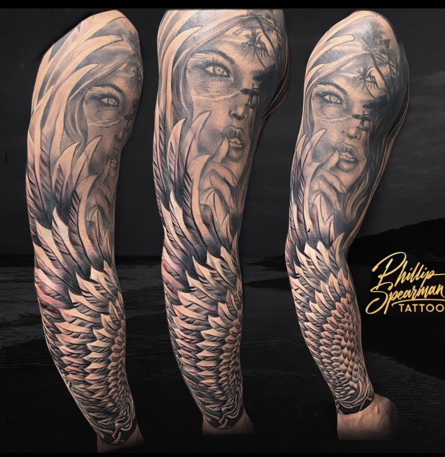 This black and grey sleeve presents a captivating narrative, featuring a woman with a finger delicately resting on her lips amidst a serene tropical landscape. The precise glint in her eyes draws you in, while a tiny surfer silhouetted against palm trees and countless layered feathers further down the arm add remarkable depth to this gracefully carried piece.